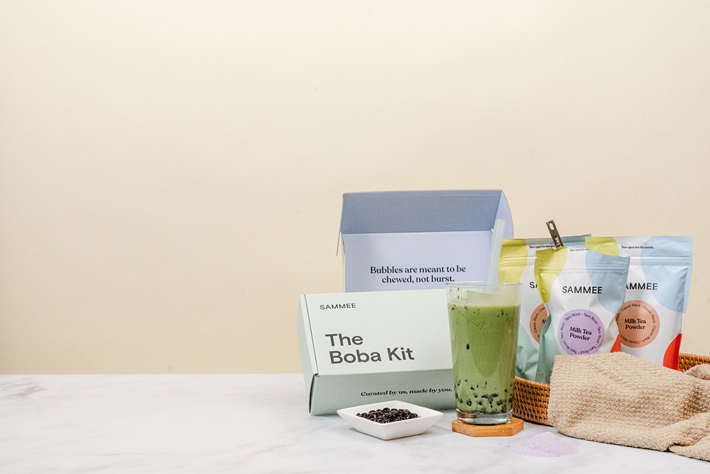 Matcha green milk tea in a clear glass on a coaster, The Boba Kit box behind it, a saucer of boba beside the glass in front of the box, and SAMMEE milk tea powders on the other side of the glass. There's also an open box of The Boba Kit in the background