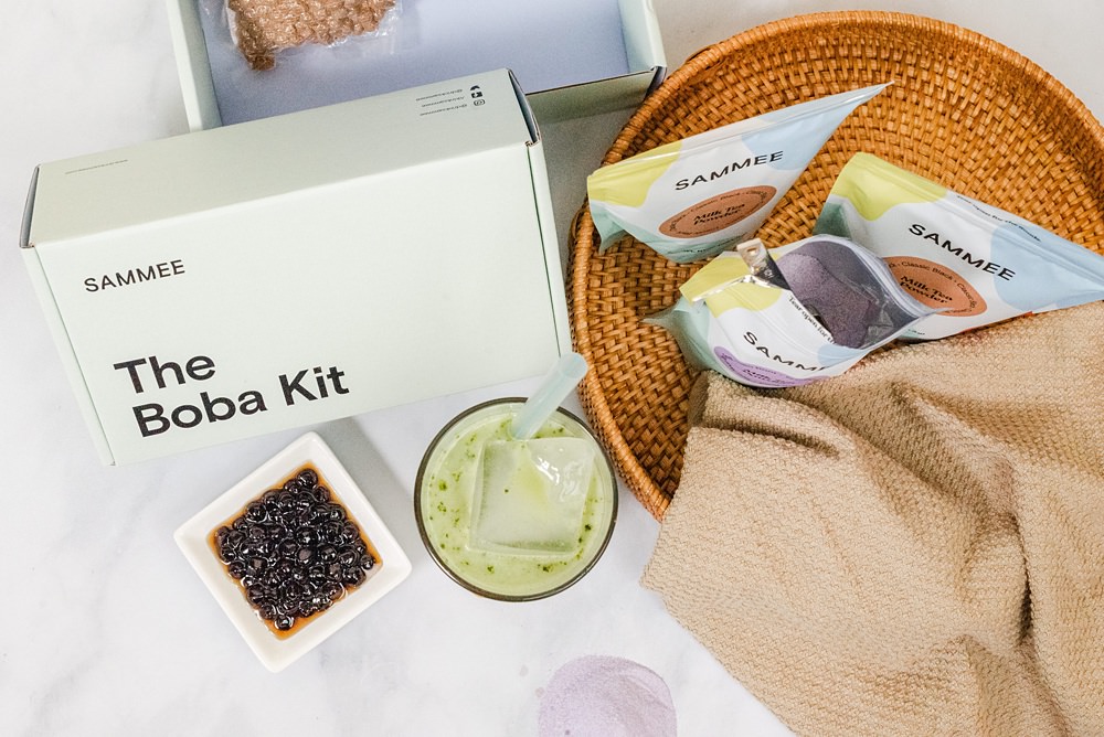 flatlay image of a matcha green milk tea in a clear glass with a block of ice and a straw, The Boba Kit box behind it, a saucer of boba beside the glass in front of the box, and SAMMEE milk tea powders, with one of them open and has a serving spoon in it, on the other side of the glass.