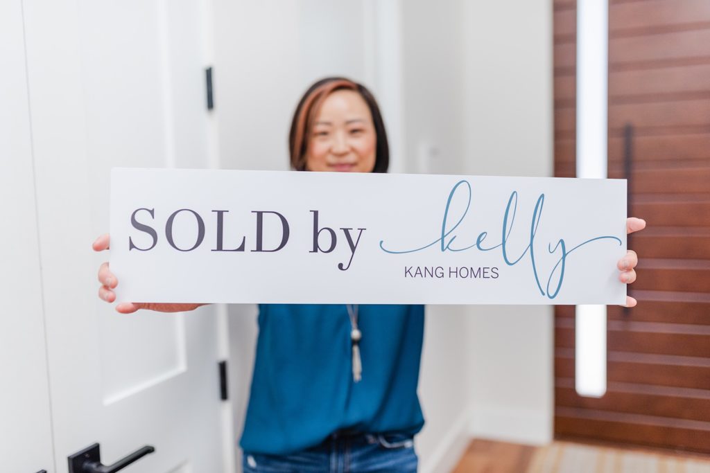 small signboard that says " SOLD by Kelly KANG HOMES" held by a woman in the background. she's standing by a white set of doors