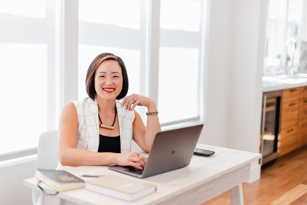smiling woman in a white vest is sitting on a white desk with an open laptop in front of her. one hand is on the laptop keyboard,