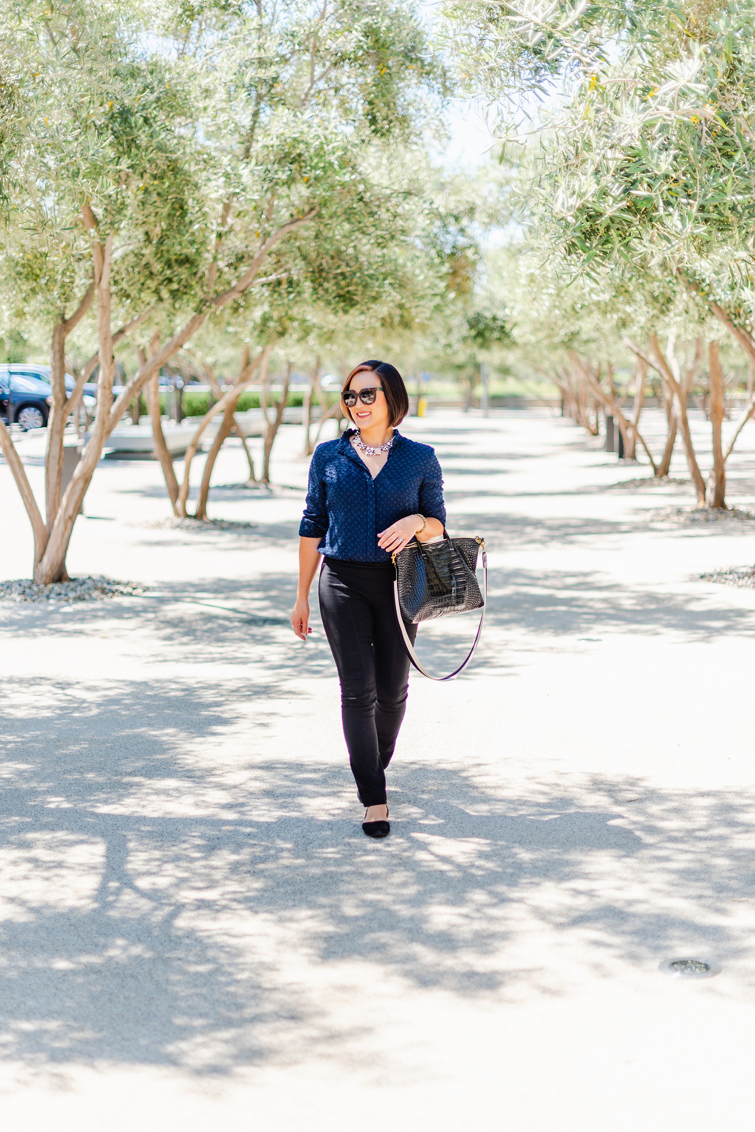 woman in blue blouse is carrying a handbag on her arm while walking between two lines of trees
