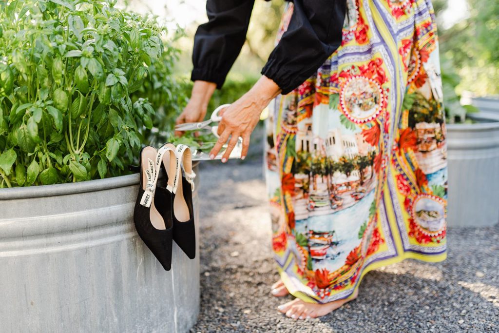 black heels hanging off the side of a trough planter with someone in a colorful, printed skirt and black long sleeves cutting some leaves while barefoot