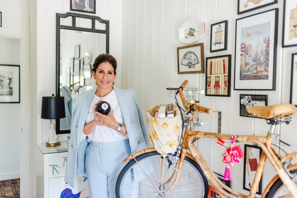 Woman in a blue suit with the jacket draped on her shoulders is holding an eight ball. there's a wooden bicycle beside her and a bunch of artworks on the white wall to her right.