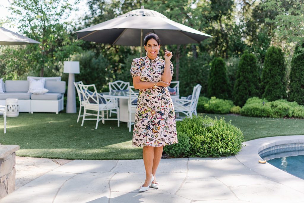 woman wearing a butterfly print collared dress is standing on a backyard with one arm loosely pointing up while the other is across her stomach holding her elbow