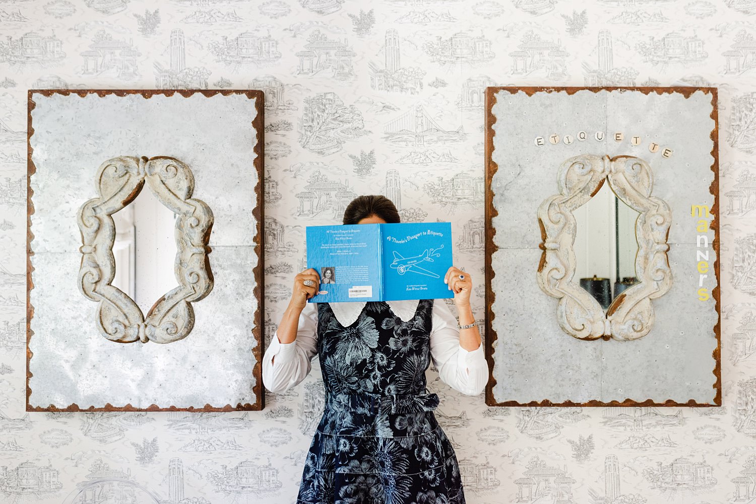 Woman in dark floral dress is holding an open book up to hide her face. She is standing between two artworks with mirrors in the center, the one on her left has the words "etiquette" and "manners" on it.