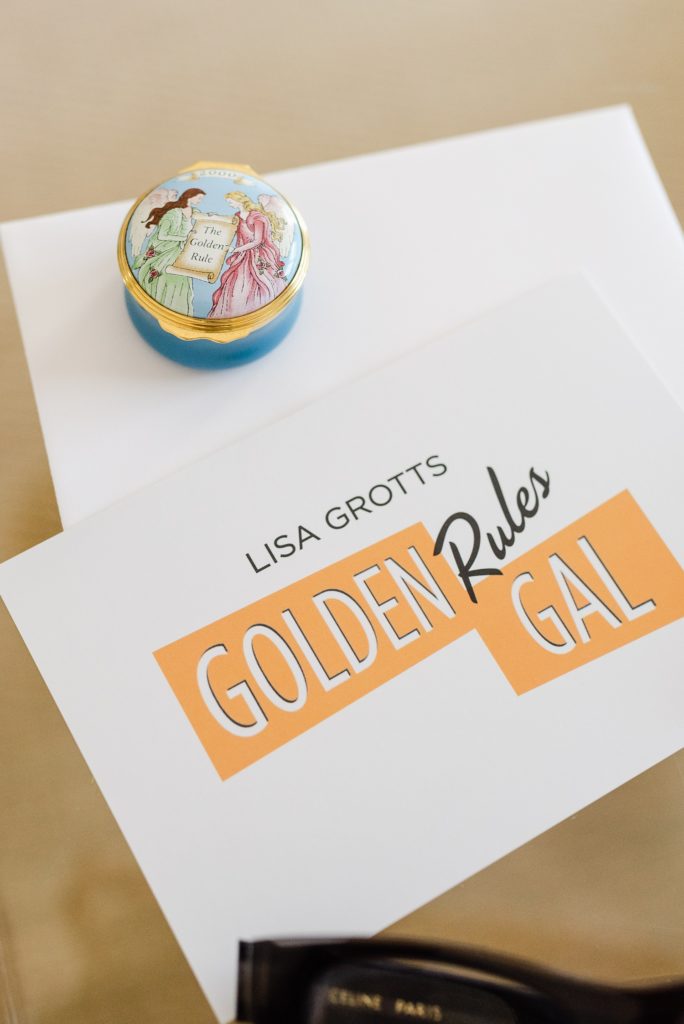 papers on a surface with "Lisa Grotts Golden Rules Gal" logo on it. There's a little trinket with illustration of two ladies holding a scroll that says "The Golden Rule" on top of the paper