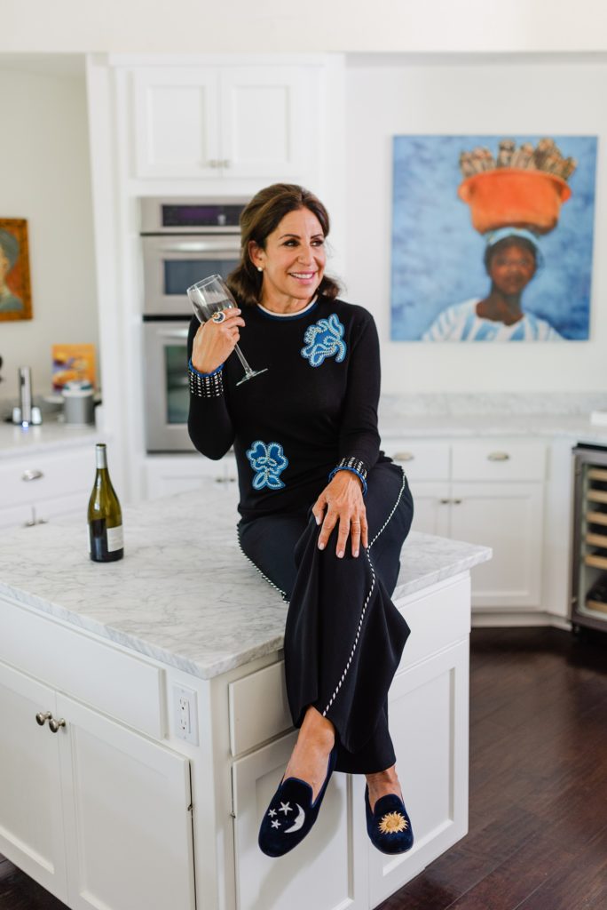 woman sitting with one knee over the other on the countertop while holding a glass of wine, the wine bottle is beside her on the counter
