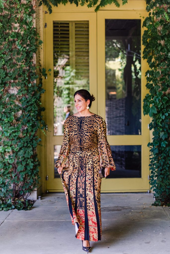 woman wearing an animal print dress is walking on pavement. there is a yellow french door behind her with shrubs growing around it's frame