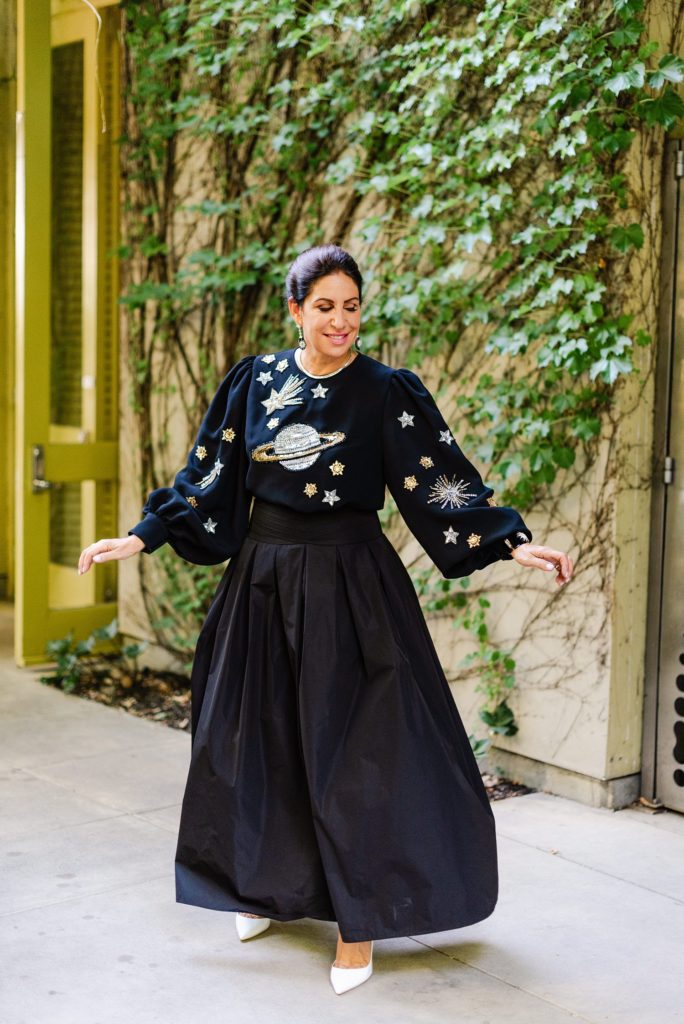 woman in long black skirt and long sleeved galaxy embroidered blouse prancing around. the wall behind her has vines crawling up on it