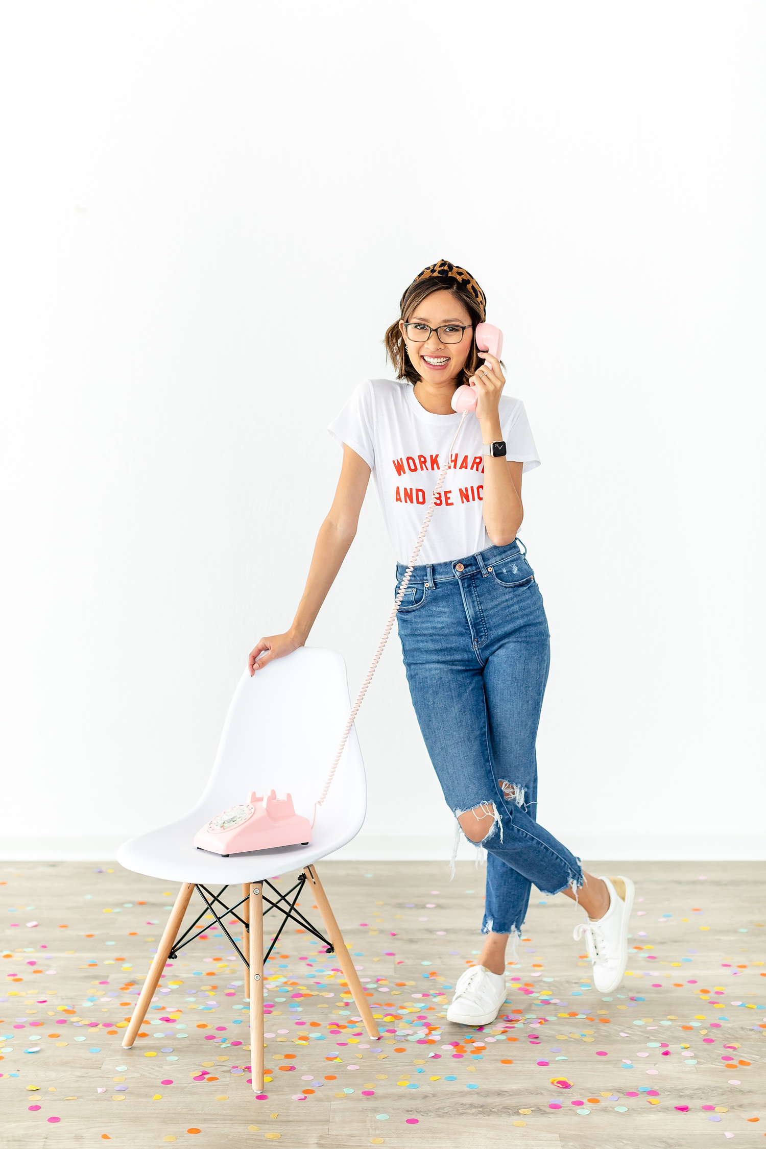 woman wearing a shirt that says "work hard be nice." She's leaning on a white chair and holding a pink retro cord phone.