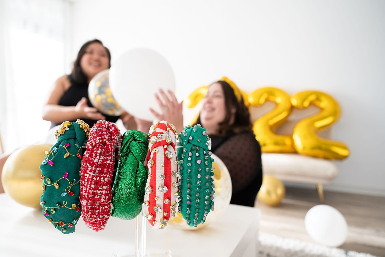 Multiple headbands on in front with colors green, red, silver green, striped red. Headbands come with different designs. At the background, there are two happy women holding a balloon and celebrating new year.