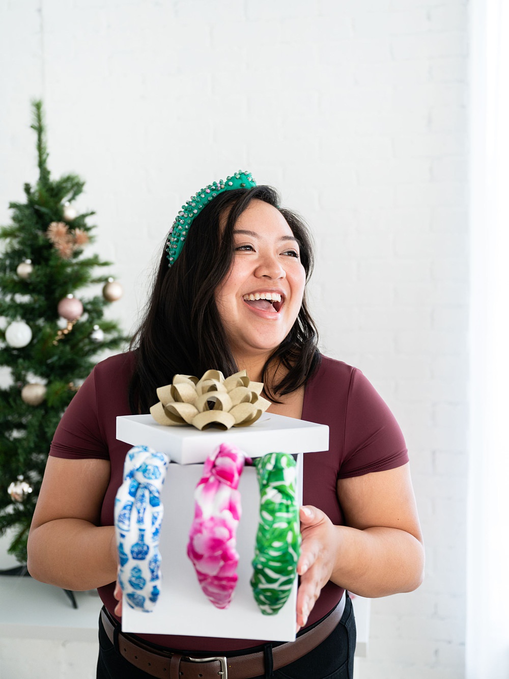 A happy woman beside the Christmas tree holding a slightly opened white gift box with three more headbands. There are blue, pink and green headbands inside the box.