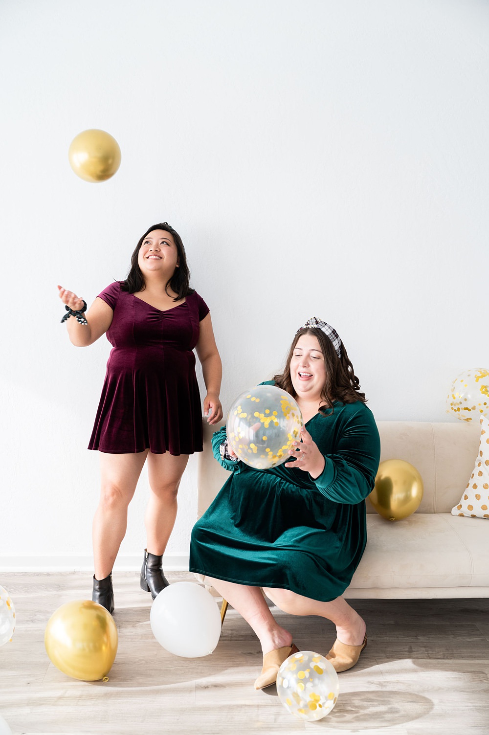 Two women holding balloons. The other woman is standing up and the other is sitting on the couch.