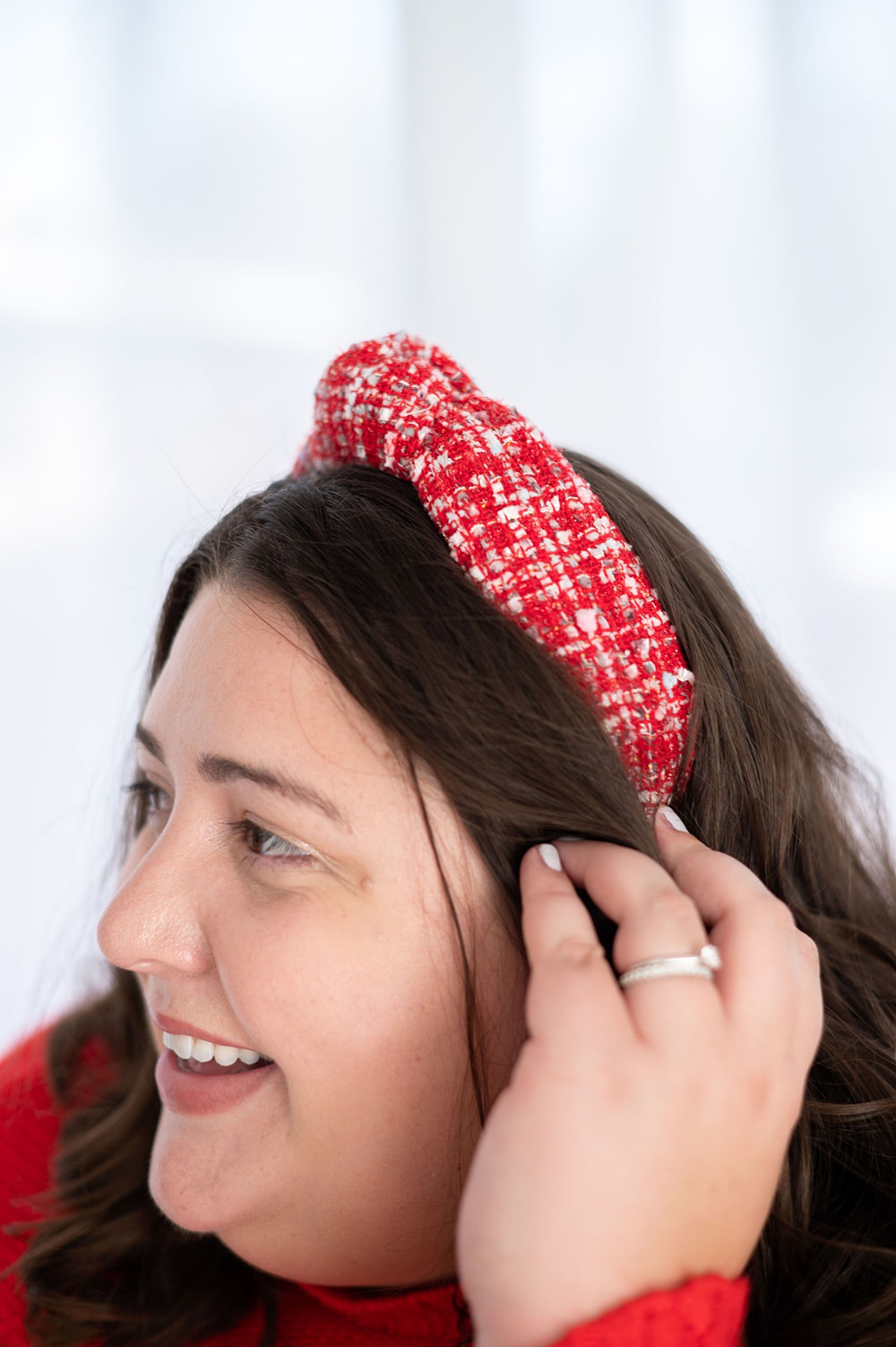 A woman wearing a red headband smiling while tucking her hair to her ears. She's looking to the left.