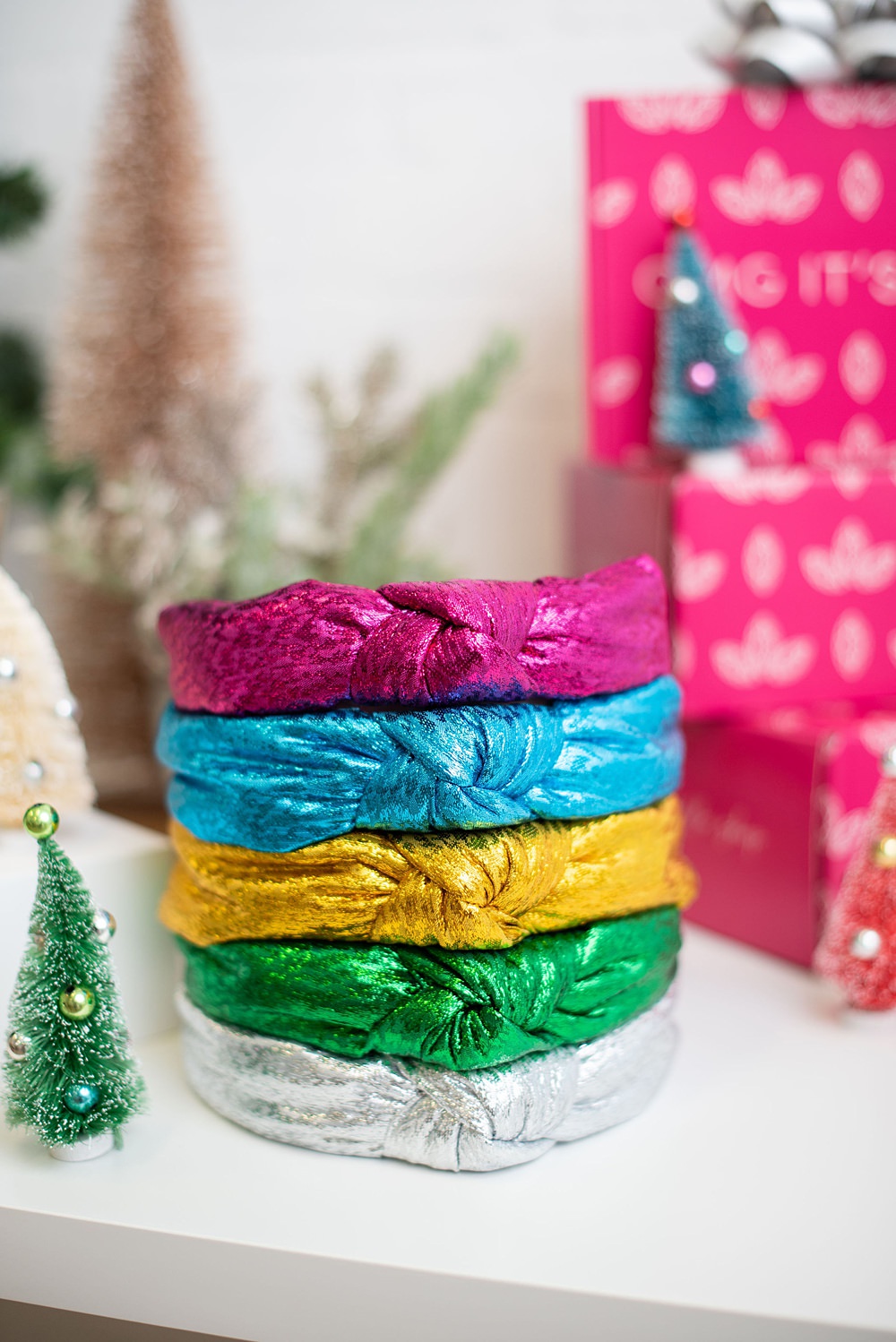 Multiple headbands on the table beside Christmas decors. There are silver, silver green, gold, silver blue and silver pink colored headbands.