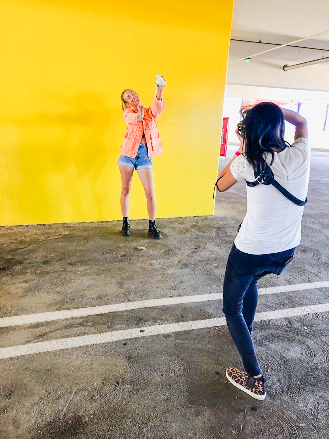 A photographer taking a picture of its  subject. The subject is a woman against the wall wearing orange jacket, a crop top and shorts.