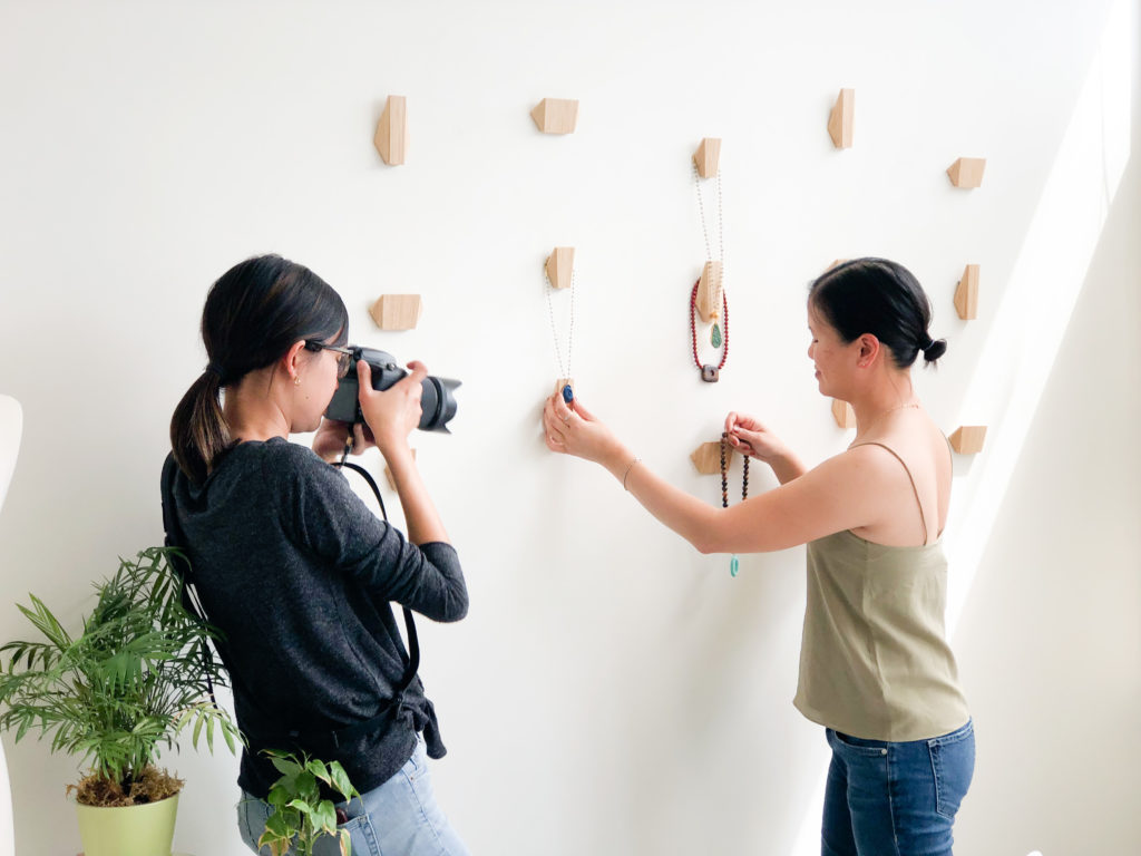 A woman photographer taking a picture of its subject who is another woman holding necklaces against the wall.