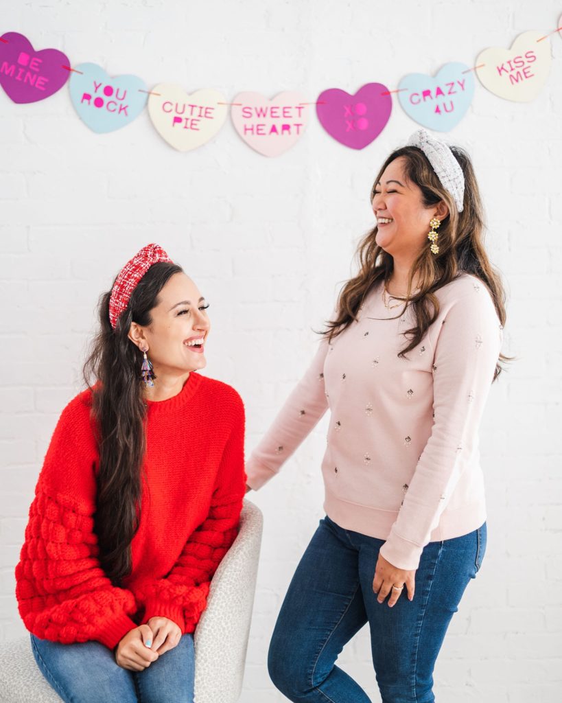 two women laughing at each other wearing headbands. There is a Valentine's Day garland hanging behind them.